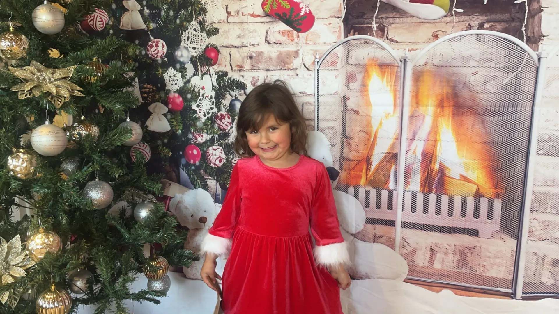 Girl in red dress smiles near Christmas tree and fireplace scene.