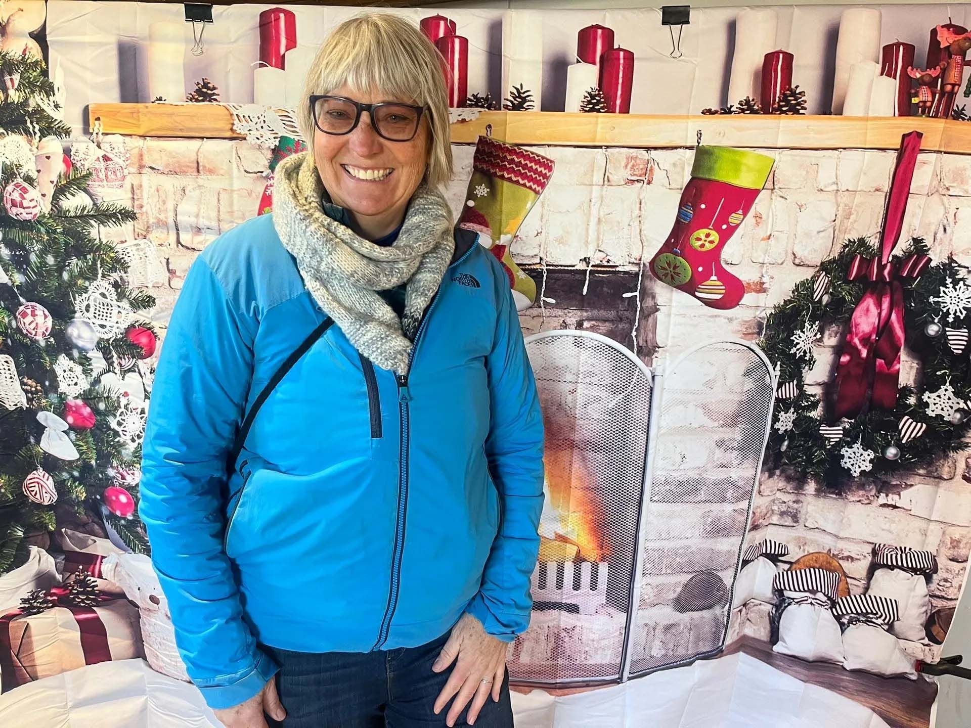 Woman in blue jacket, scarf, and glasses smiles in front of a Christmas backdrop.