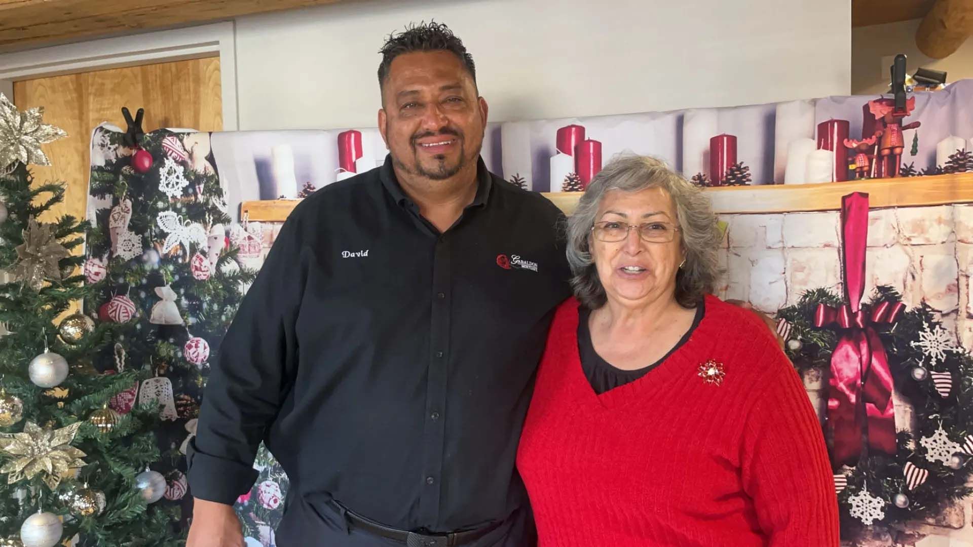 Man in black shirt and woman in red sweater stand together in front of a Christmas backdrop.