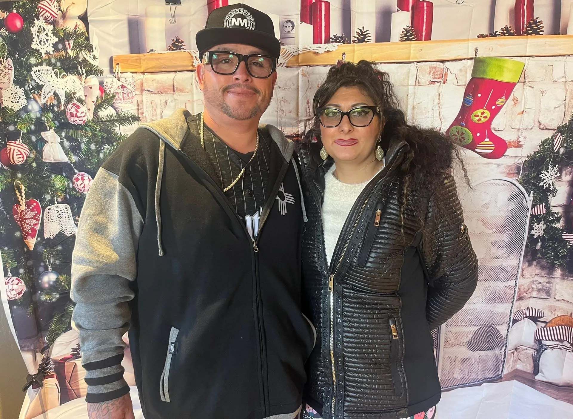 Couple posing in front of a Christmas backdrop. They wear glasses and winter jackets.