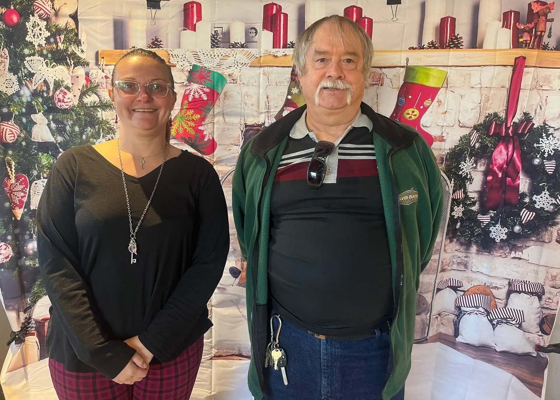 Woman and man posing in front of Christmas decorations; man wearing a green jacket.