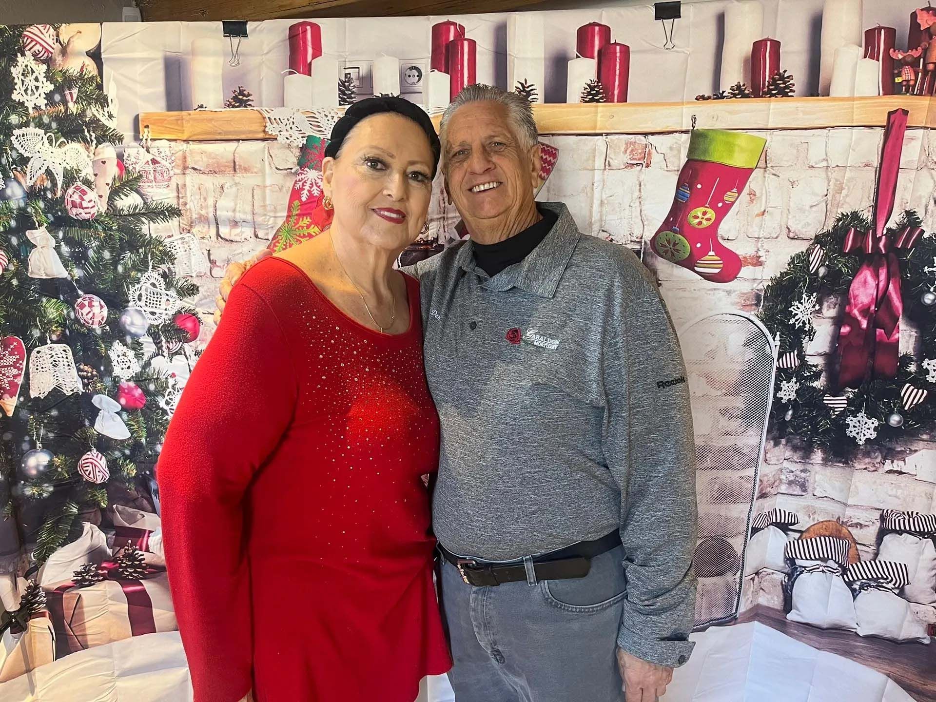 A couple poses in front of a Christmas-themed backdrop. Woman wears red, man in gray. Smiling.