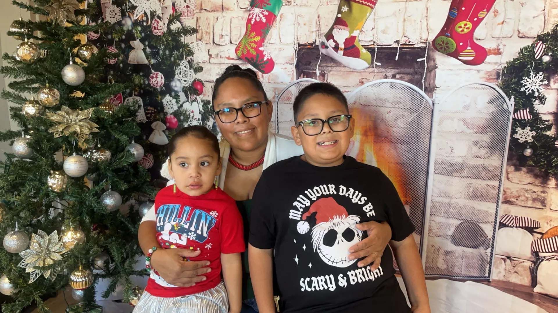 Woman and two children pose by Christmas tree and fireplace backdrop.