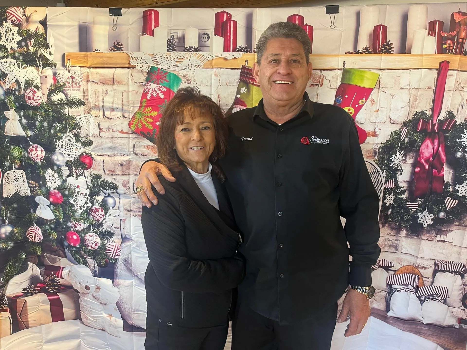 Two people, a woman and a man, pose in front of a Christmas backdrop. They are smiling.