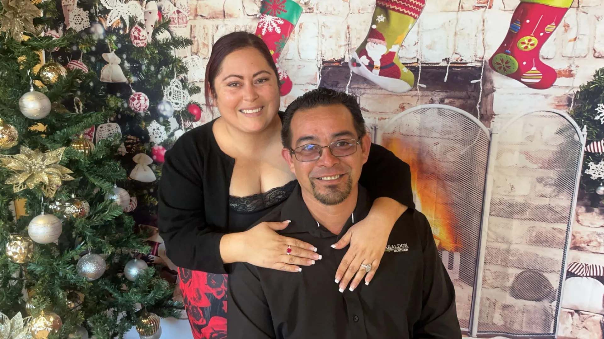 Couple posing, woman's arms around man, in front of Christmas tree and fireplace backdrop; smiling.