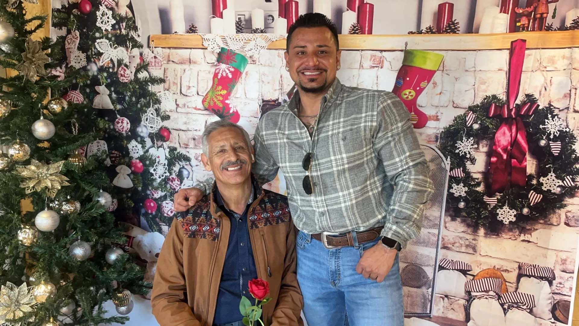 Two men pose in front of a Christmas backdrop. One man wears a brown jacket and holds a rose; the other wears a plaid shirt and smiles.