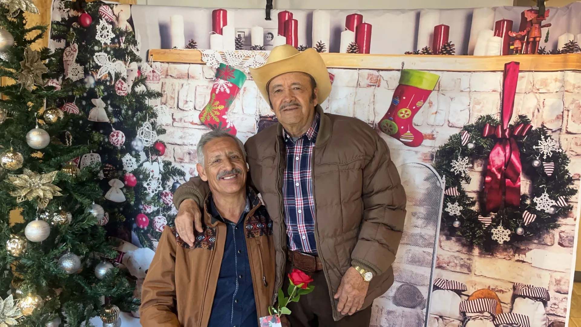 Two men pose in front of a Christmas tree and backdrop with stockings and a wreath. One wears a cowboy hat.