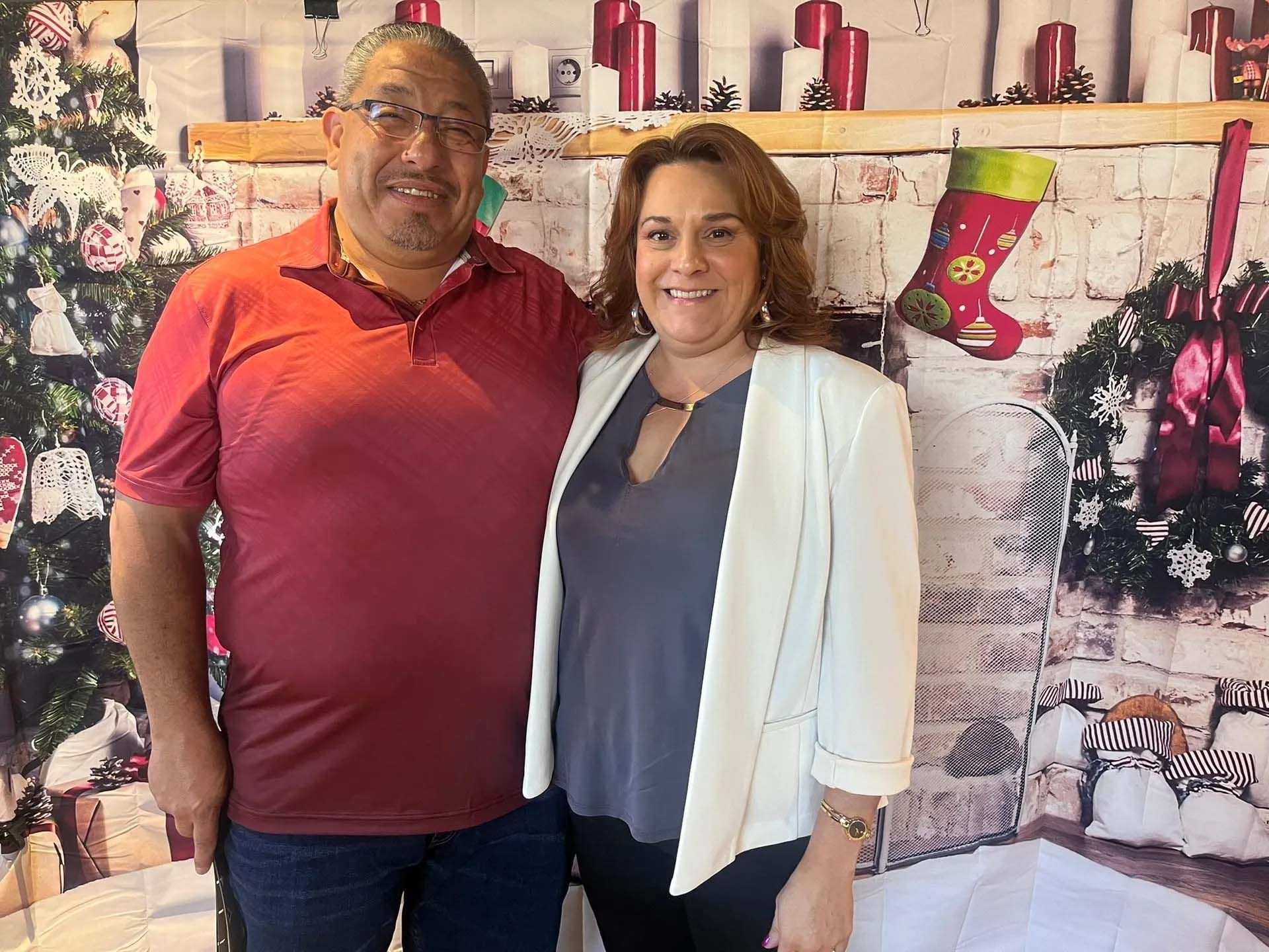 Man in red shirt and woman in white jacket pose in front of a Christmas backdrop.