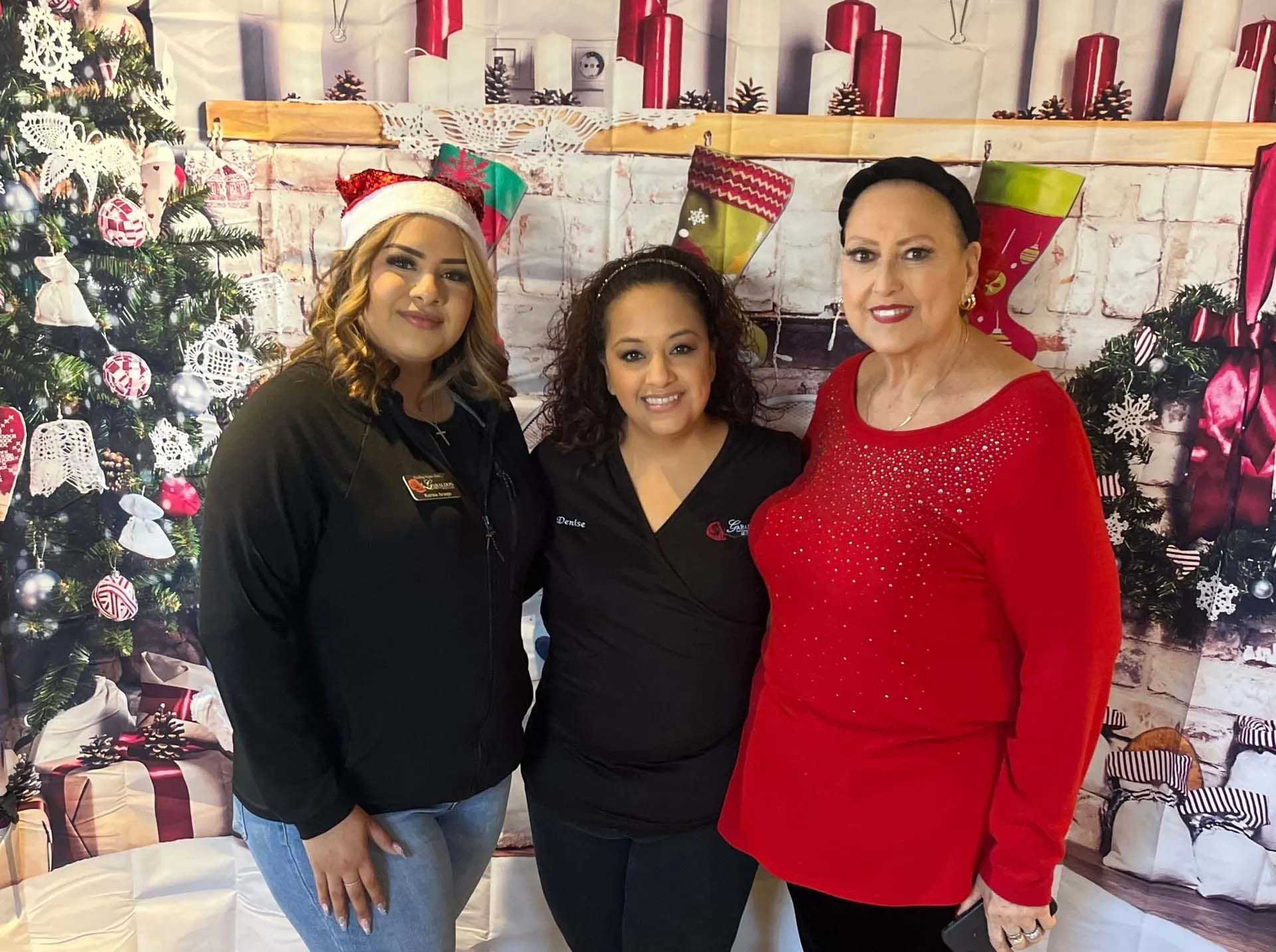 Three women smiling, posing in front of a Christmas-themed backdrop. One wears a Santa hat, another a red sweater.