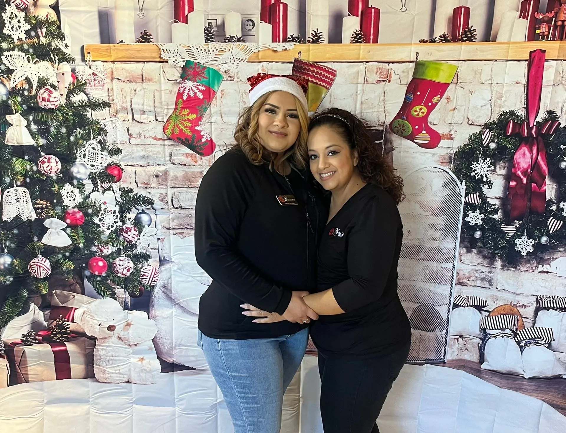 Two people smiling in front of a Christmas backdrop. One wears a Santa hat, holding the other's arm.