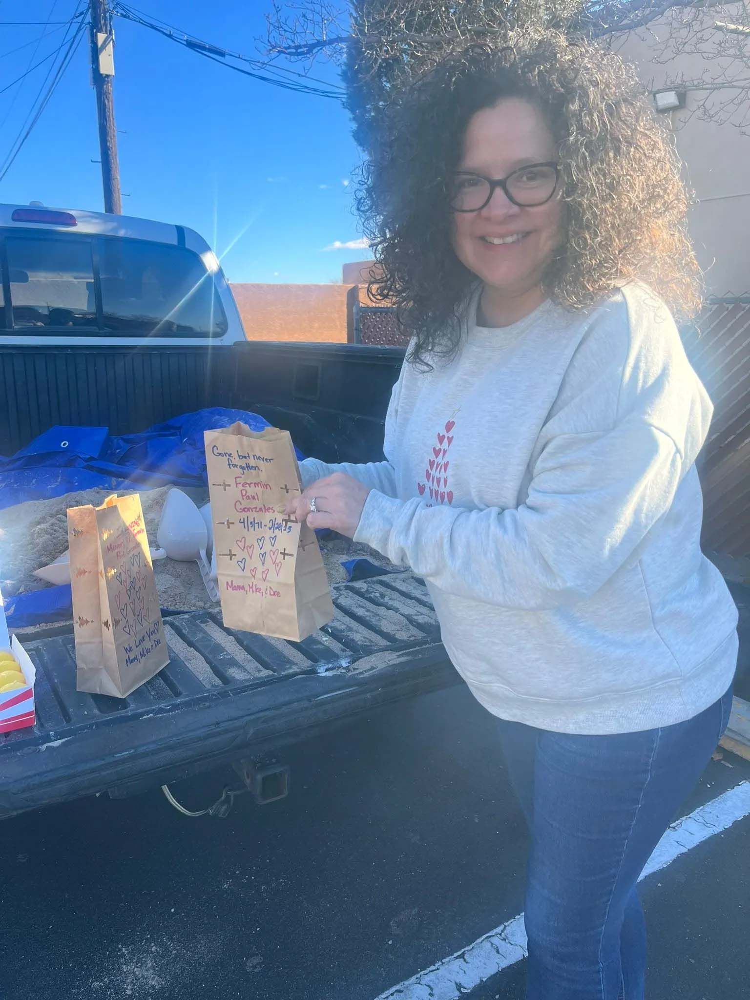 Woman in gray sweatshirt smiles, holding a brown paper bag in truck bed with others. Sunny outdoor setting.