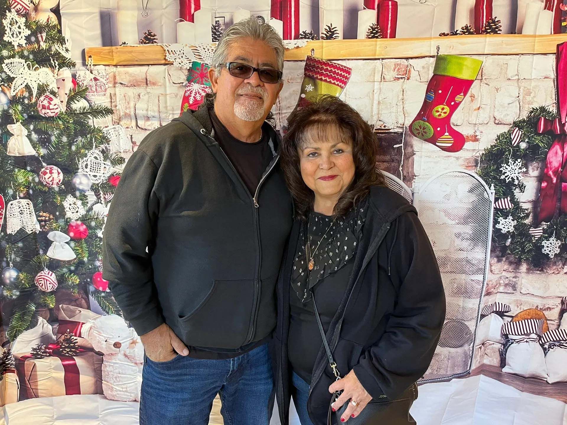 Couple in front of a Christmas backdrop with tree and stockings. Man wears sunglasses, woman smiles.