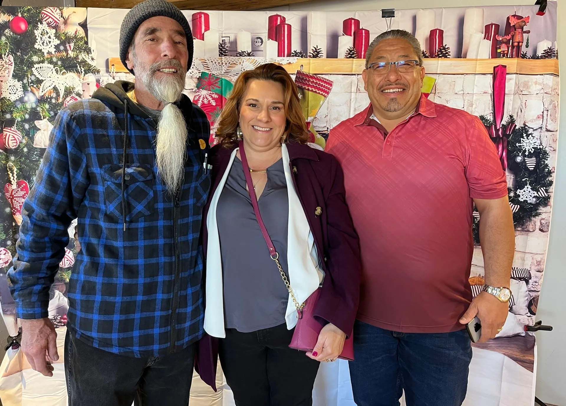 Three people posing for a photo in front of a Christmas backdrop. Woman in middle, men flanking her.