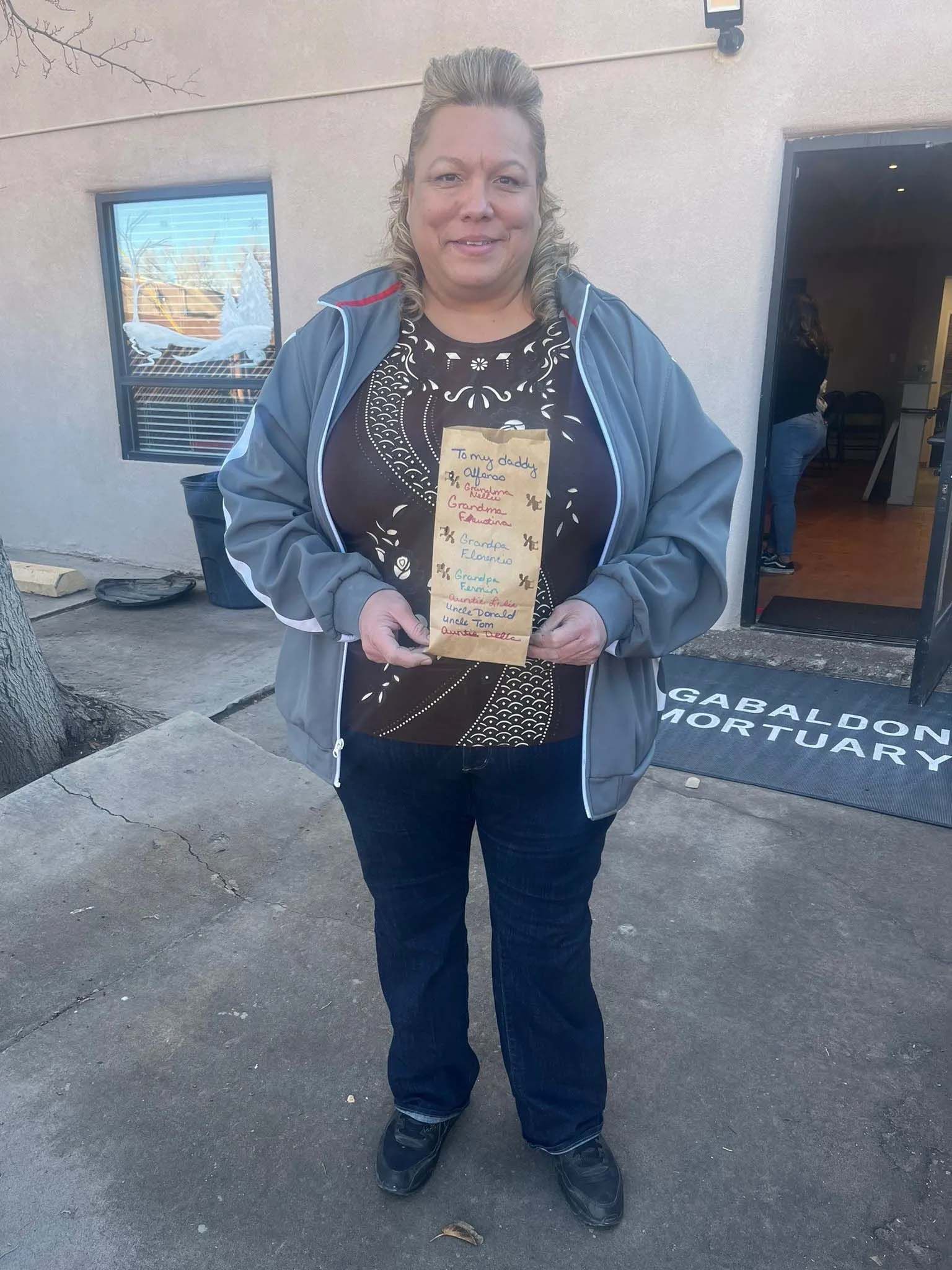 Woman in front of building holding paper, wearing a gray jacket and jeans.