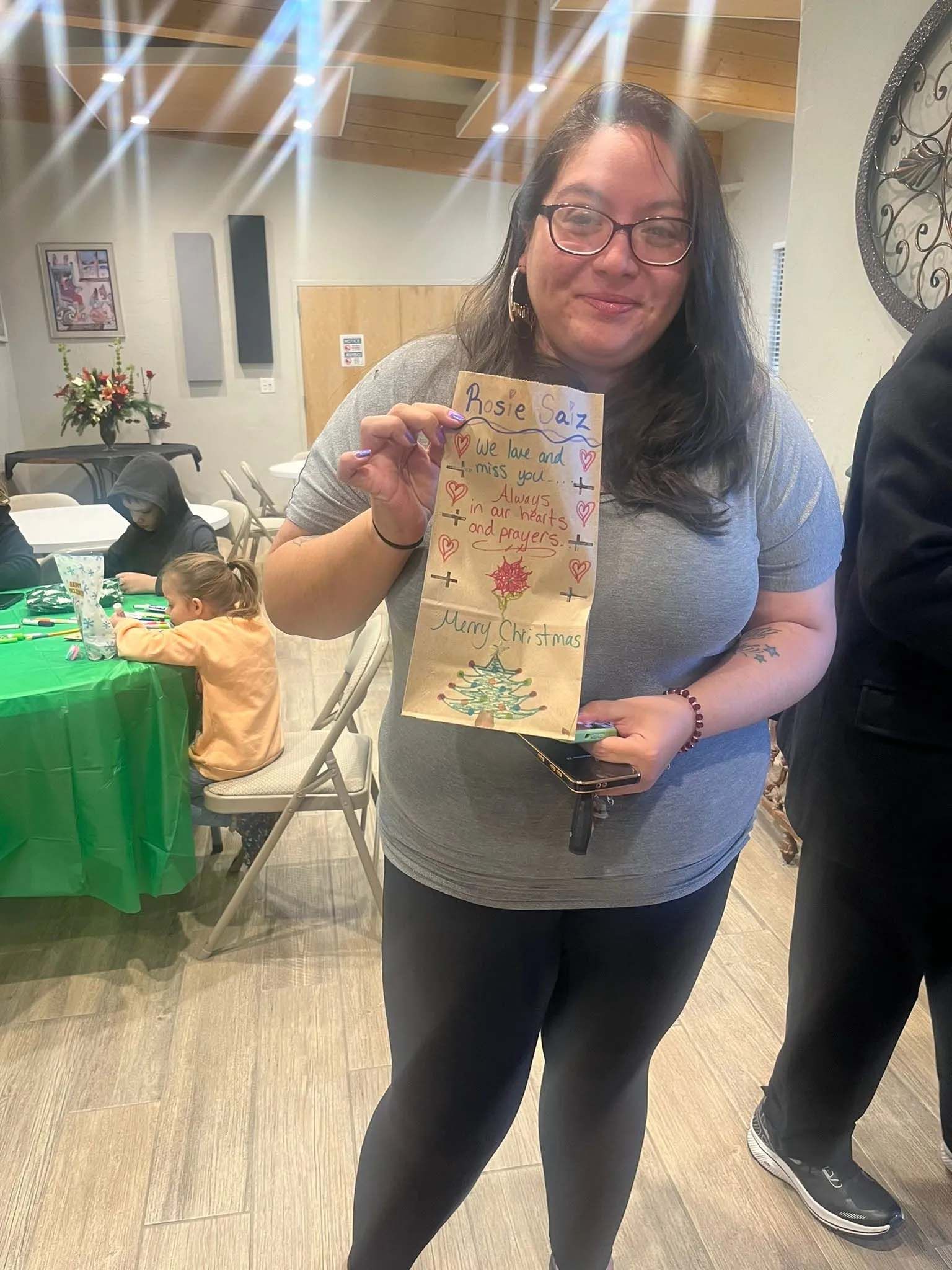 Woman holds up decorated paper bag. Indoor setting with party decorations and a child at a table.