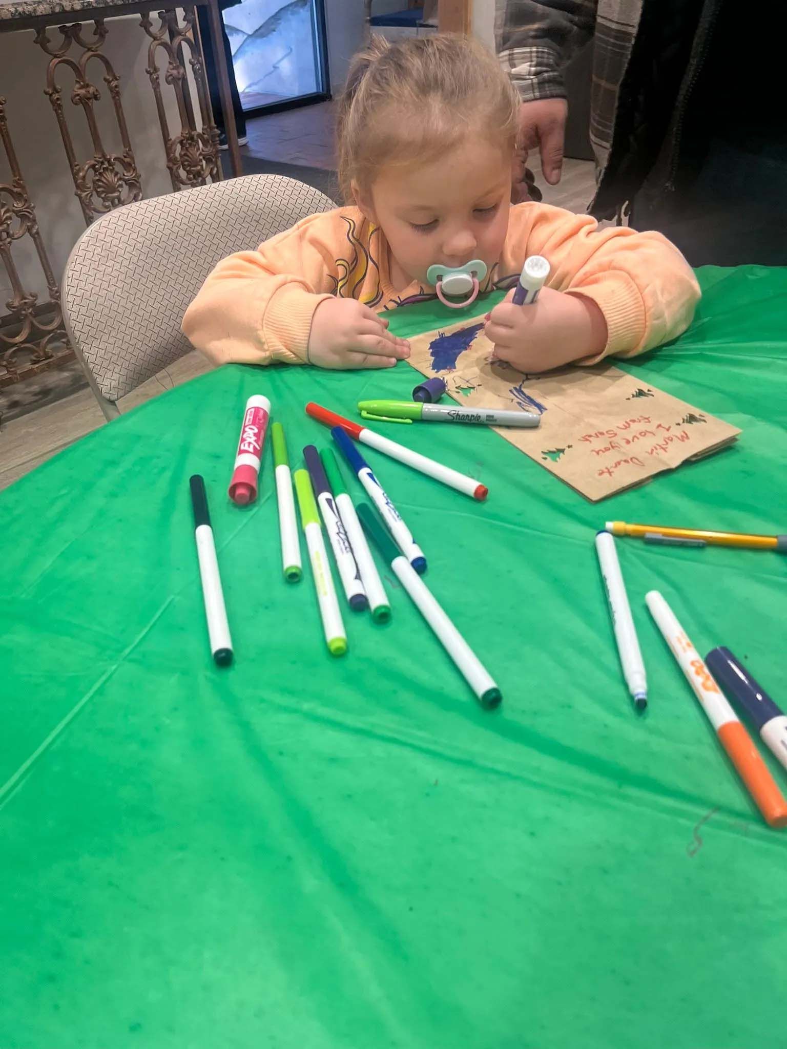 Young child coloring with markers on a green surface, holding a pacifier.