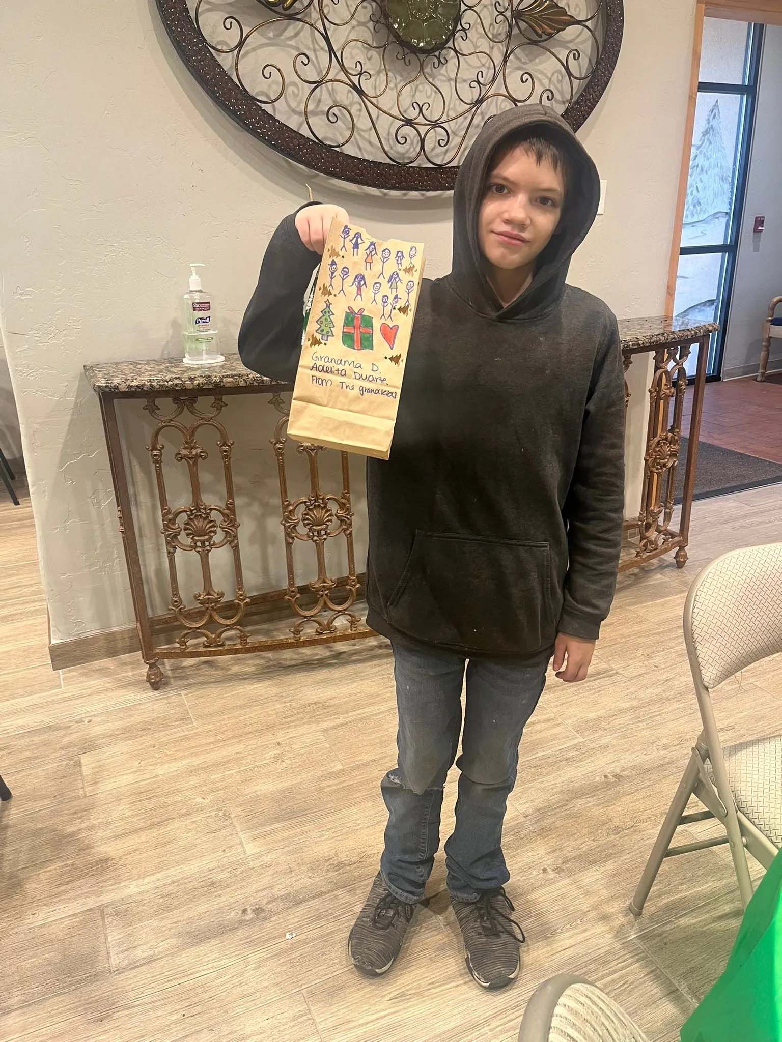 Person holding decorated paper bag, wearing a black hoodie and jeans, standing indoors.