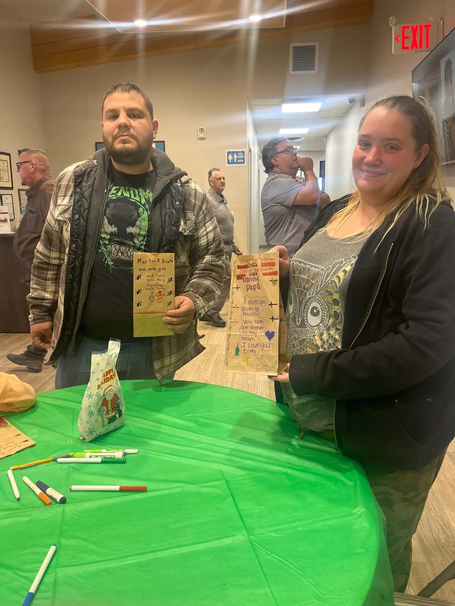 Two people stand at a table, holding decorated cardboard projects. Green table cloth. Other people in the background.