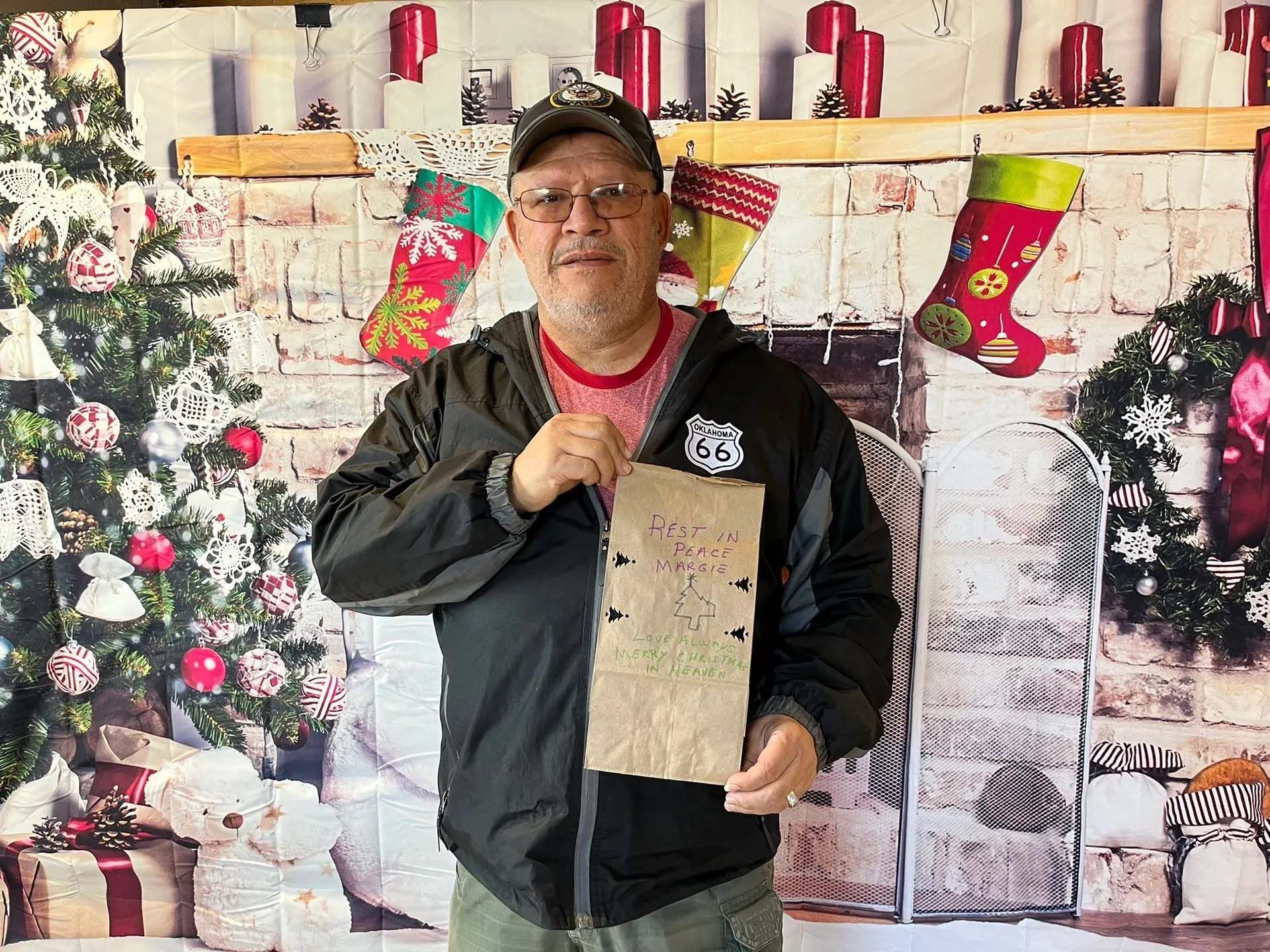 Man holding a paper with markings, standing in front of a Christmas-themed backdrop with a fireplace, tree, and stockings.