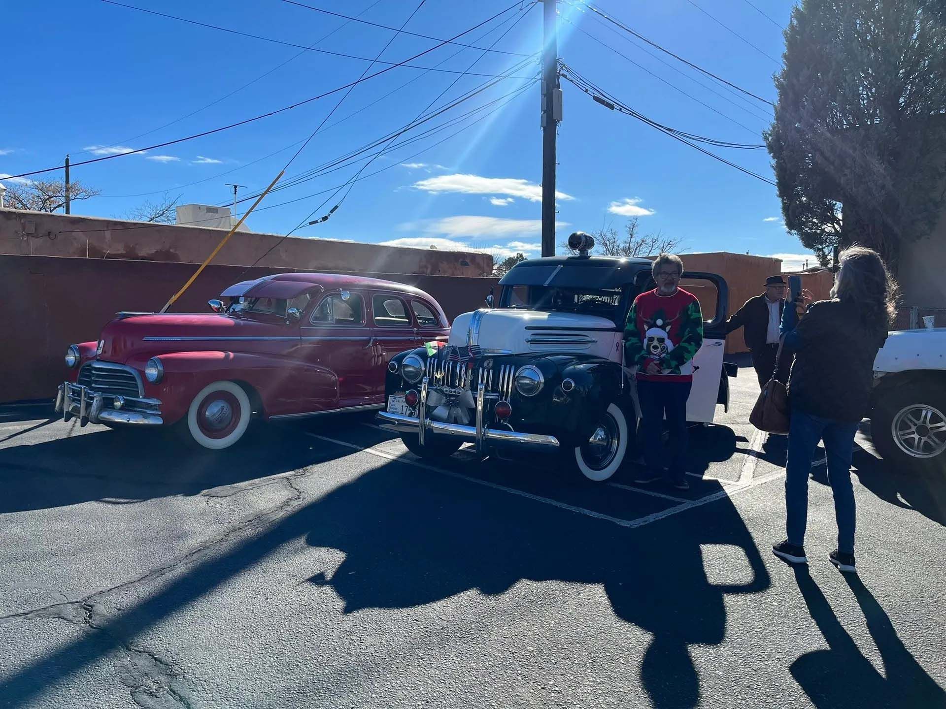 Two vintage cars parked outside; people stand near the cars on a sunny day.