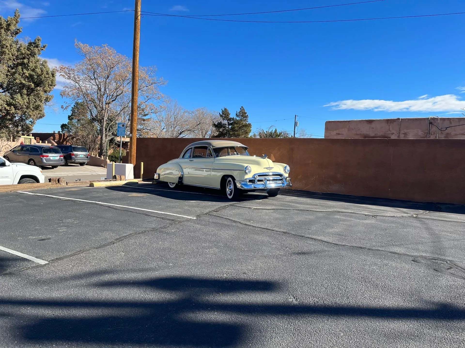 Yellow classic car parked in an asphalt lot on a sunny day. A brown wall is behind the car.