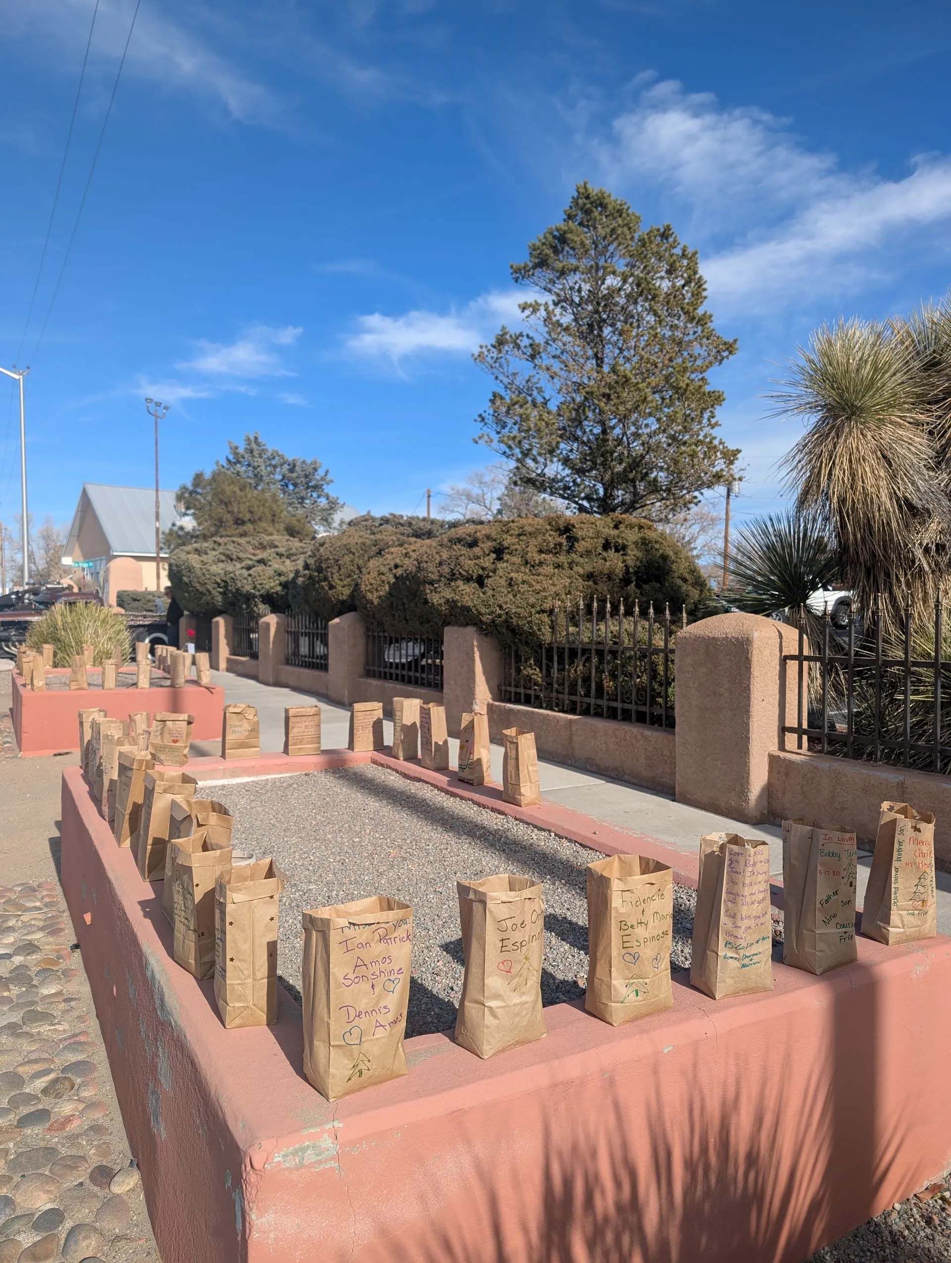 Red-walled garden beds with lit brown paper bags, sidewalk, building, trees, and blue sky.