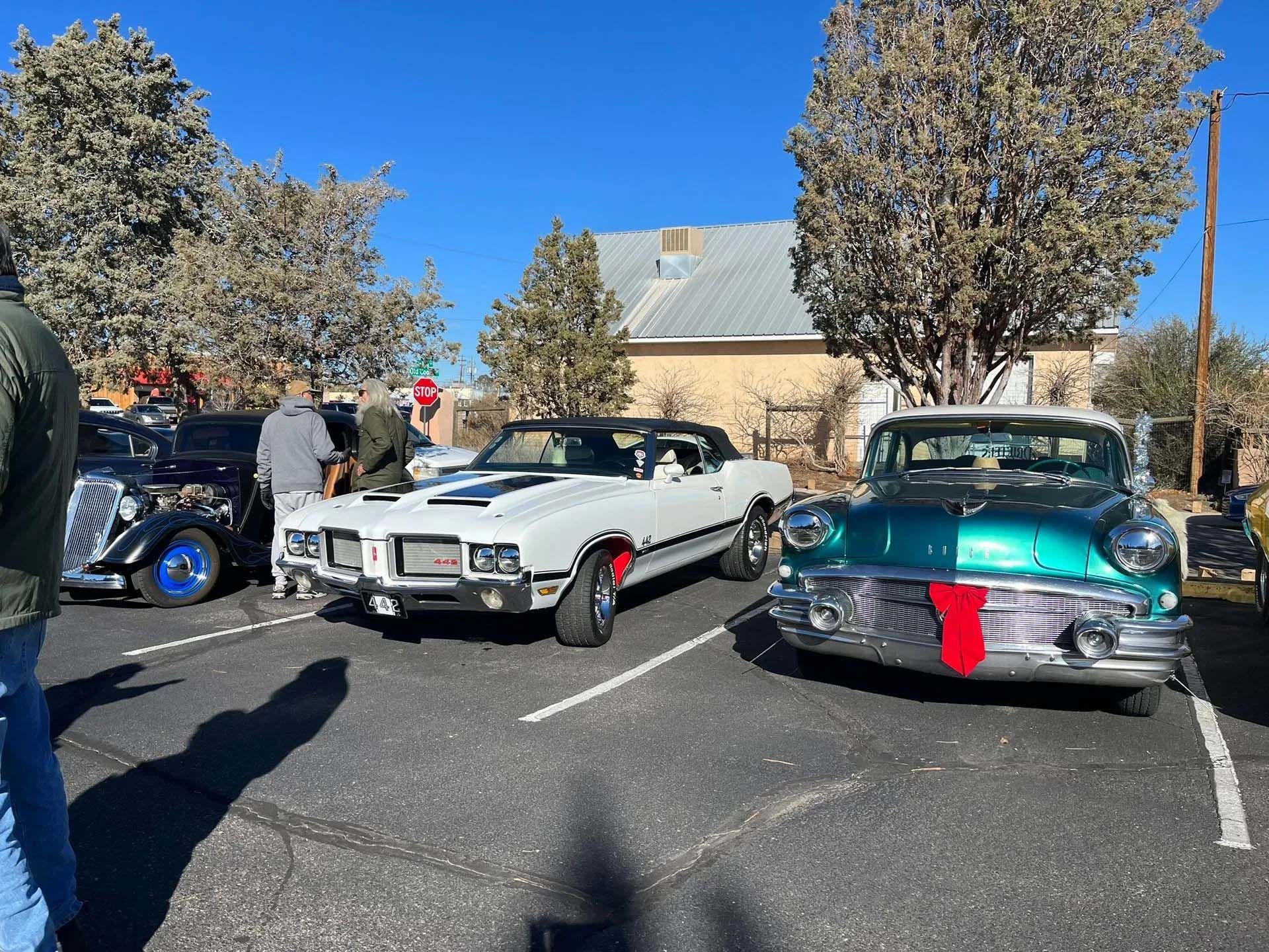 Classic cars parked outdoors: white convertible, teal car with red garland, and black car with blue wheels.