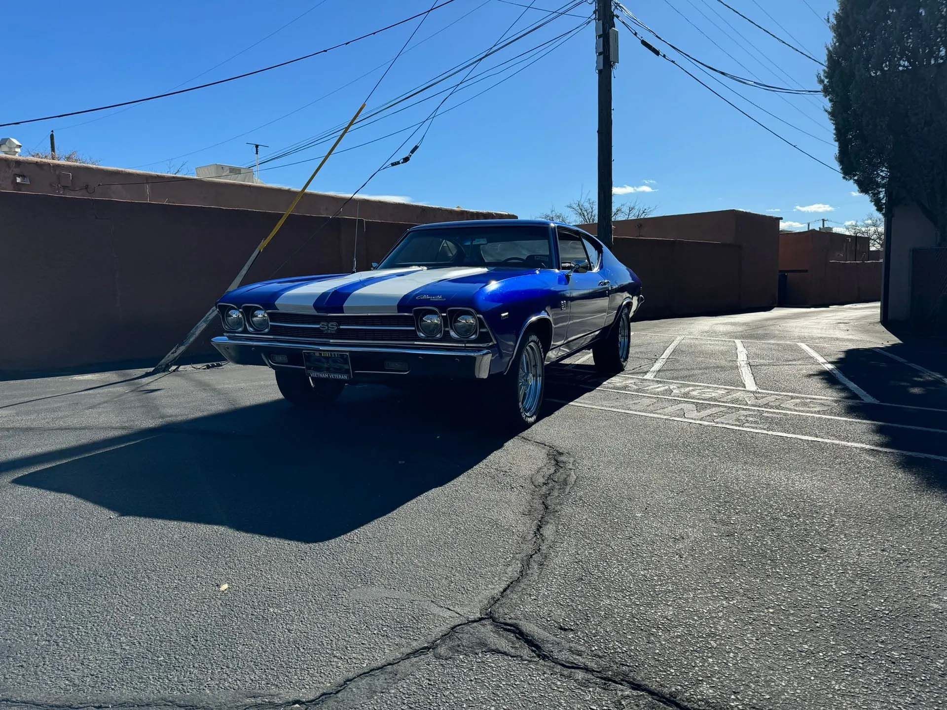 Blue classic car with white stripes parked on a paved alley on a sunny day.