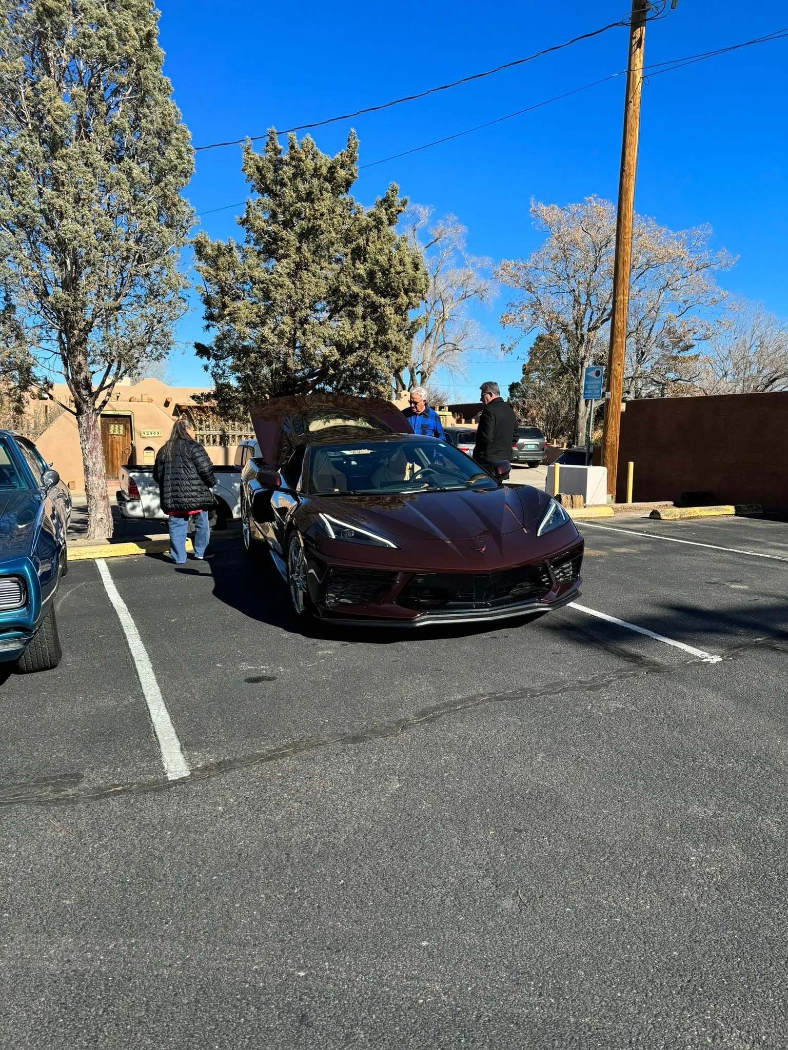 Burgundy sports car in a parking lot, hood open, people nearby. Bright blue sky, trees, and tan buildings.
