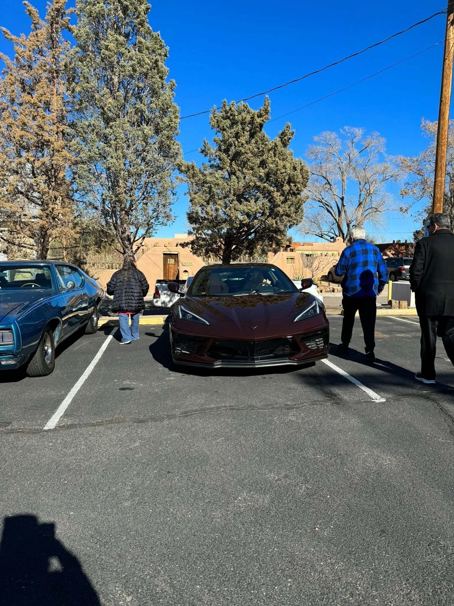 Dark red sports car parked between two other cars, people standing nearby.