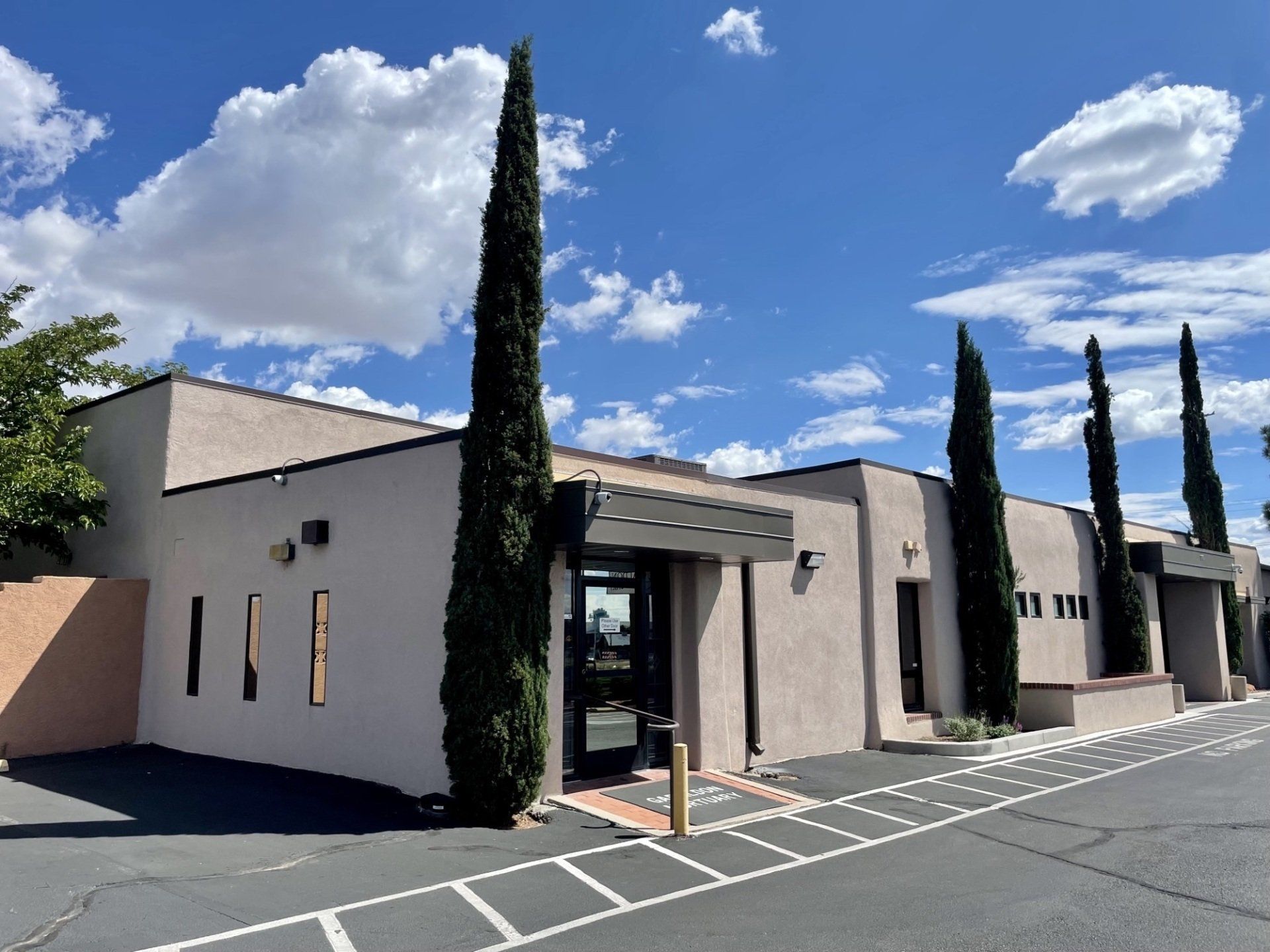 Tan building with tall, thin trees in front; blue sky with clouds.