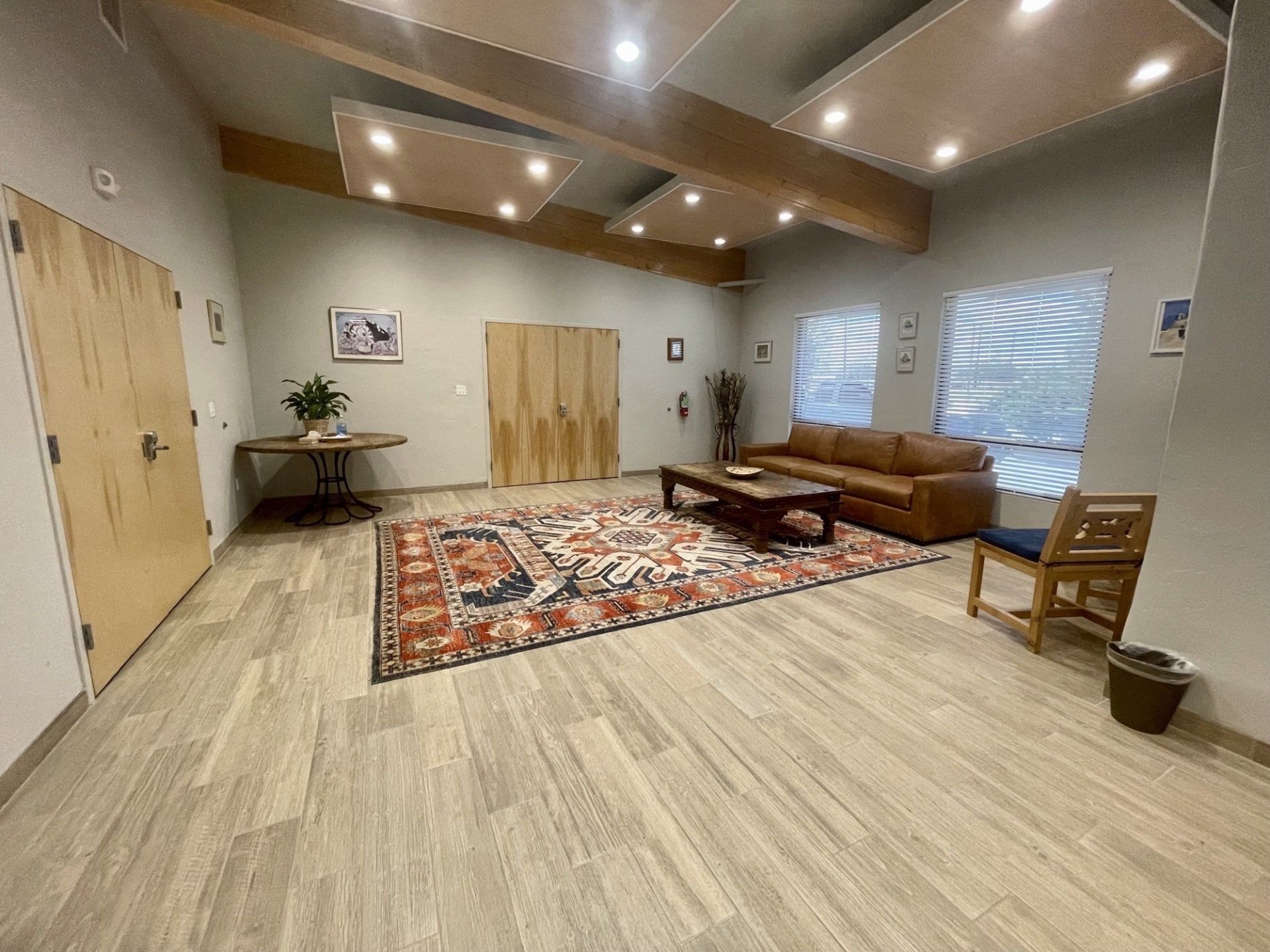 Spacious room with a brown leather couch, area rug, wooden doors and ceiling beams, and large windows.