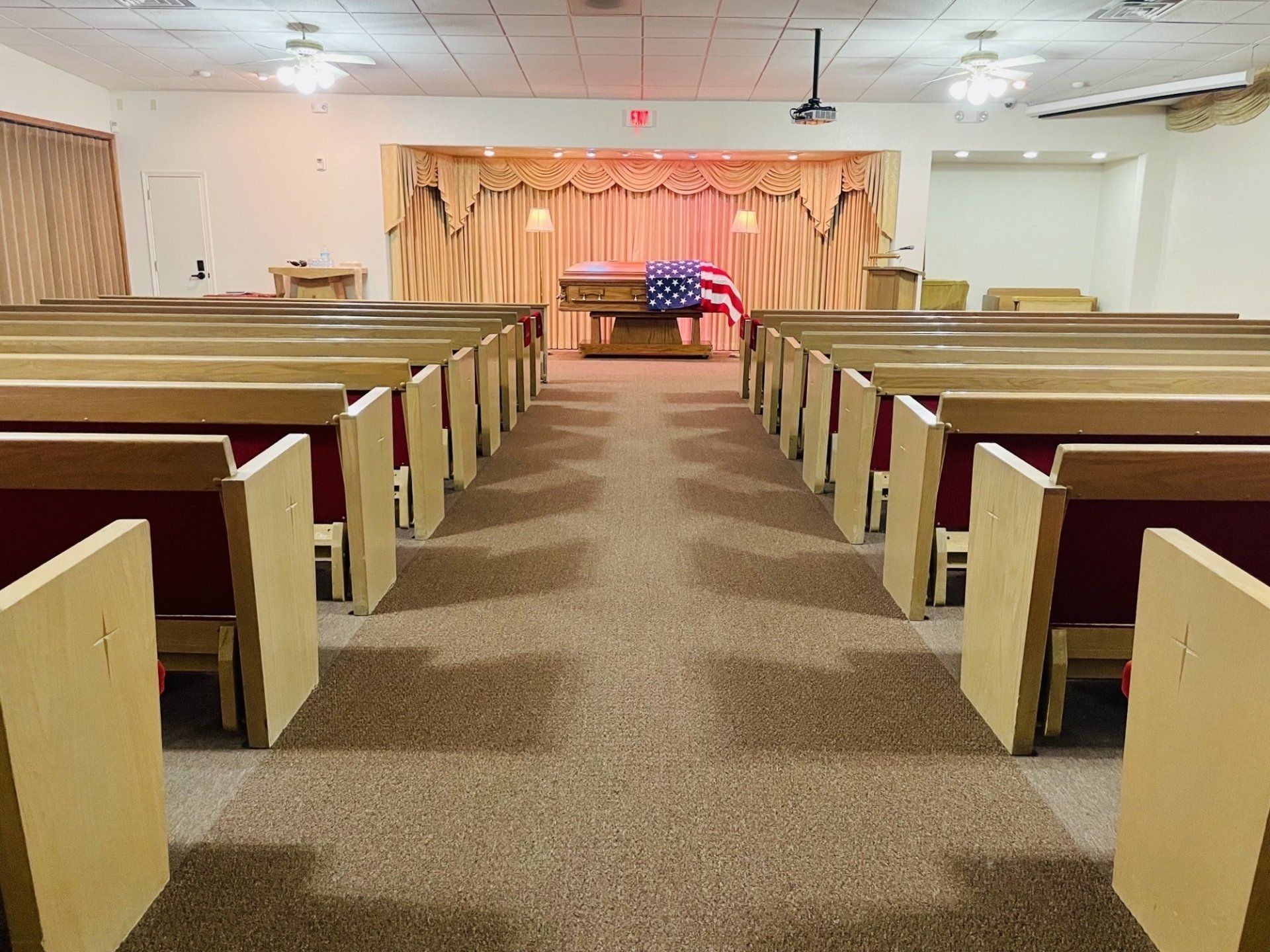 Funeral home interior with a casket draped in an American flag centered at the front, rows of empty pews.