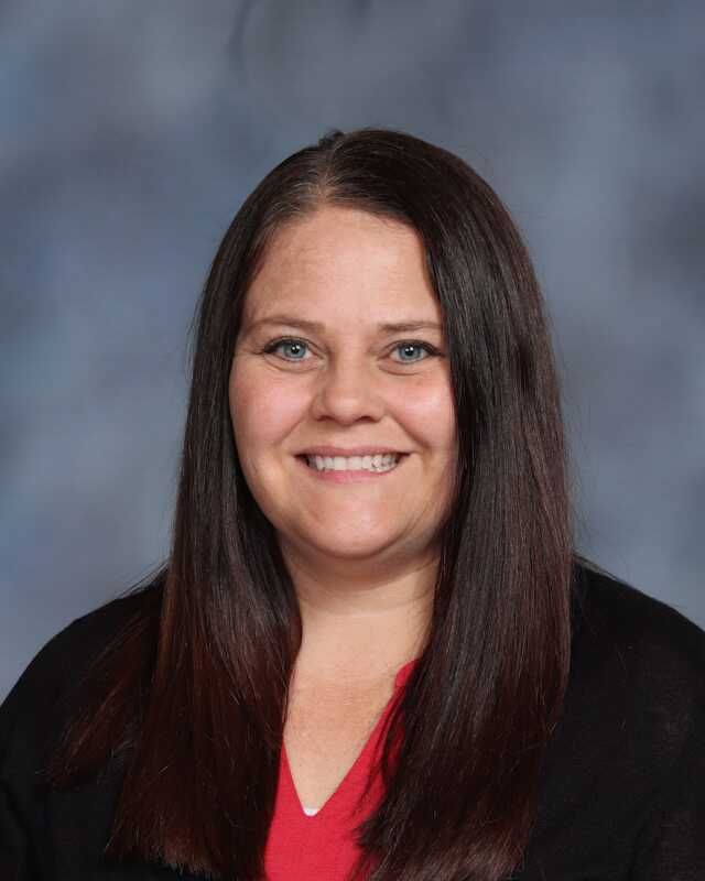 A woman in a red shirt and black jacket is smiling for a picture.