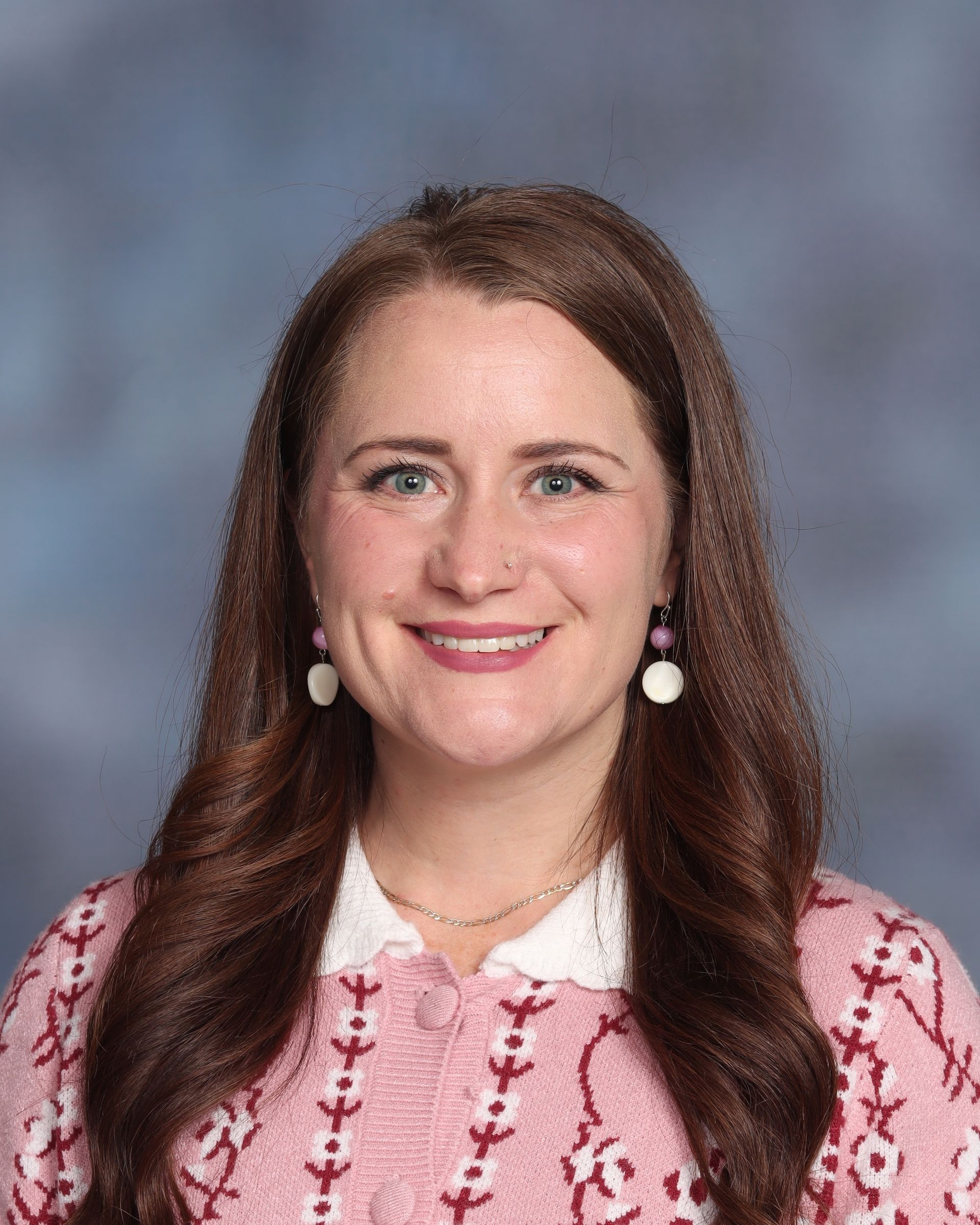 A woman with long brown hair is smiling for the camera while wearing a white shirt and earrings.