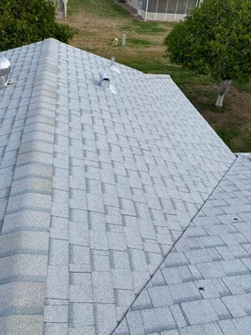 Gray shingle roof, angled view. Several vents present.  Green grass and trees in background.