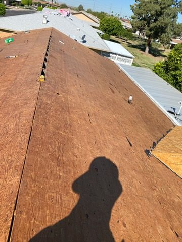 Rooftop showing brown wood panels, visible shadow, other roofs and trees in background.