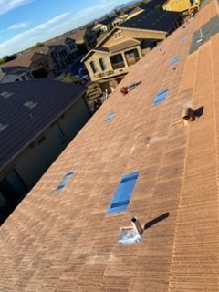 Overhead view of a brown shingled roof with skylights and chimneys, in a residential neighborhood.
