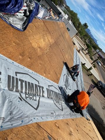 Two roofers installing flashing on a new roof. One in orange safety gear. Sunny outdoor setting.