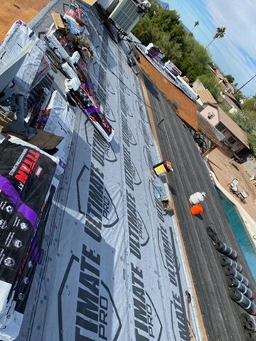 Roof under construction, with new underlayment being installed. Rolls of shingles, tools, and a pool in the background.