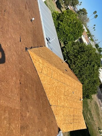 Partially roofed house, light brown plywood with exposed seams. Trees and houses in the background on a sunny day.
