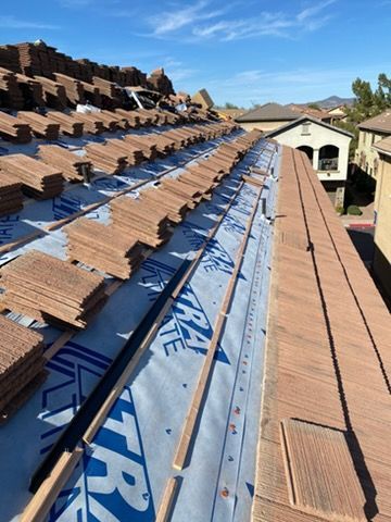Rooftop being constructed with brown tiles, blue underlayment, and wooden beams on a sunny day.