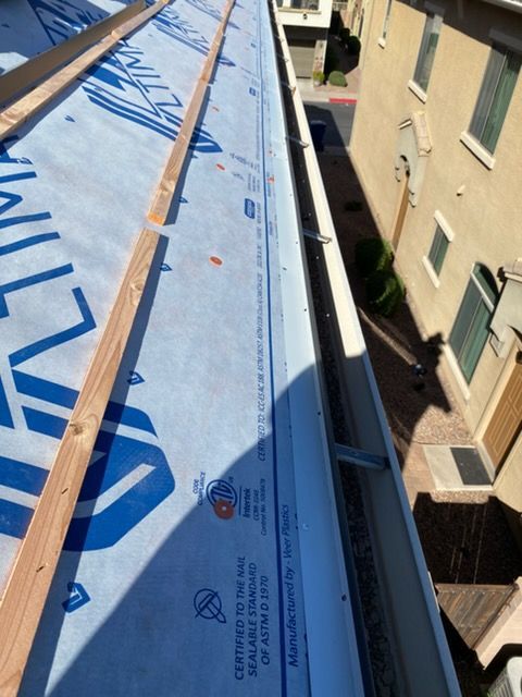 Roof under construction; blue underlayment, wooden beams, white gutter, view from above.