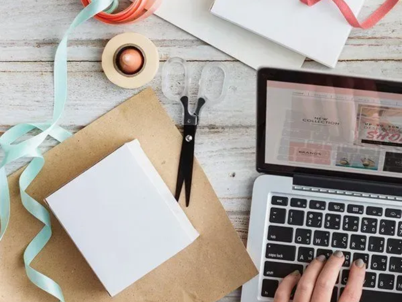 Laptop and craft supplies: Person typing on laptop near scissors, tape, ribbon, and paper.