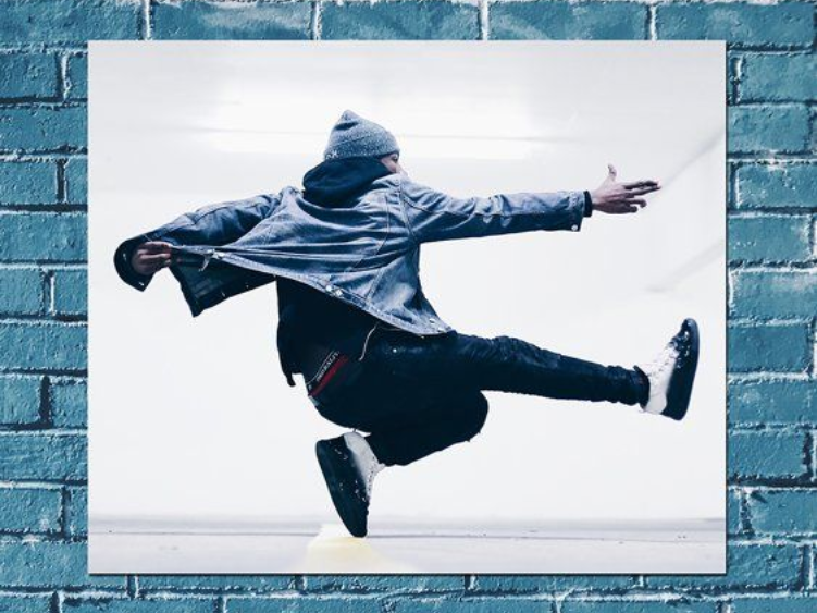 Une personne en l'air exécutant un mouvement de danse, veste en jean, bonnet et pantalon foncé.