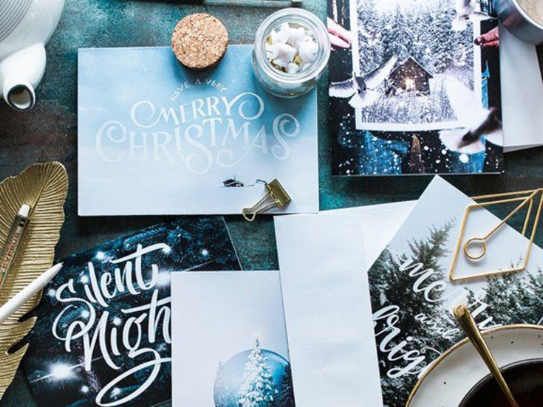 Christmas cards and stationery arranged on a dark surface; includes a teapot, gold feather, and small treats.