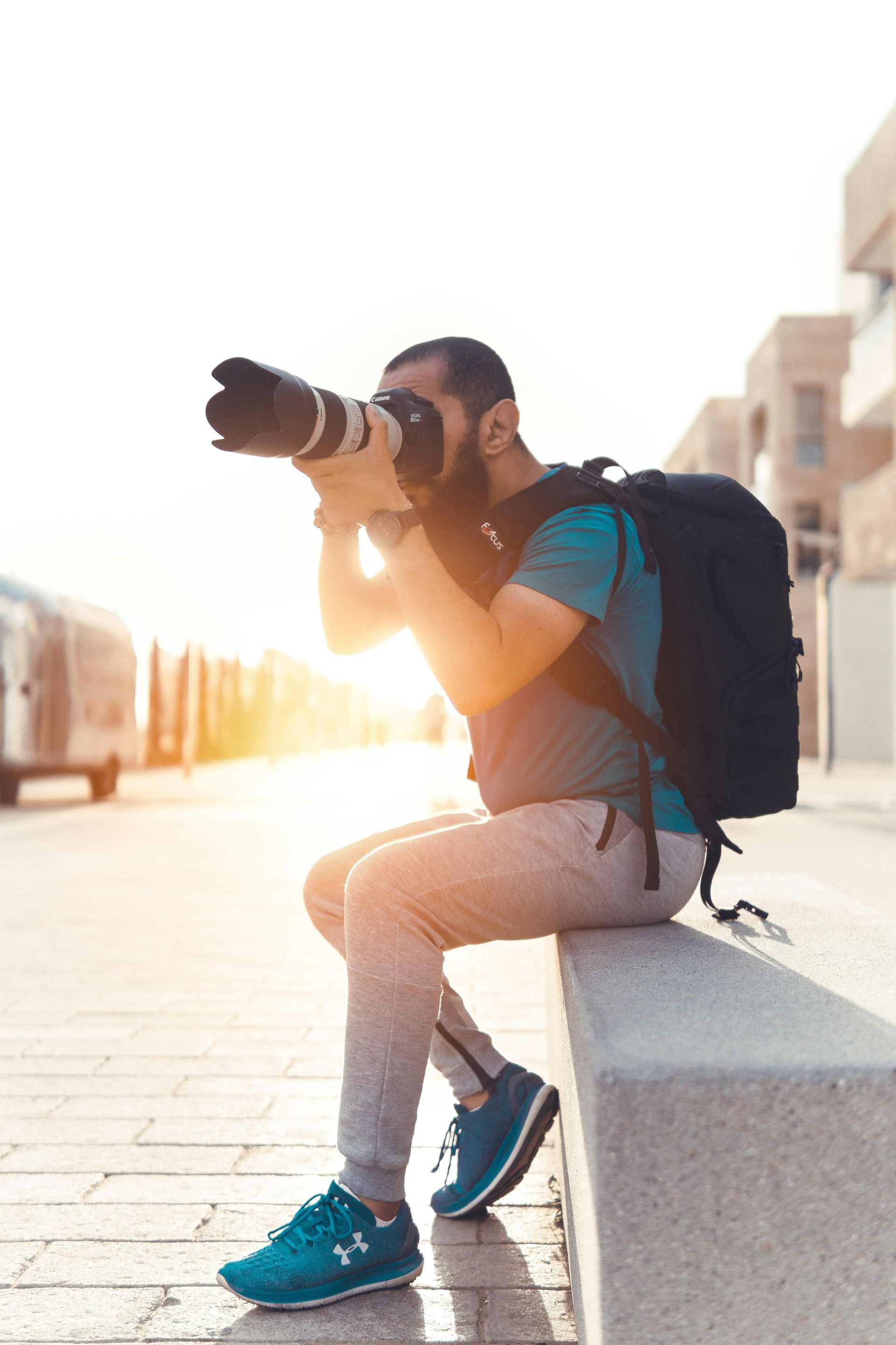 Un homme avec un appareil photo, un sac à dos, une chemise turquoise et un pantalon gris, assis, pointe l'objectif vers la lumière du soleil.
