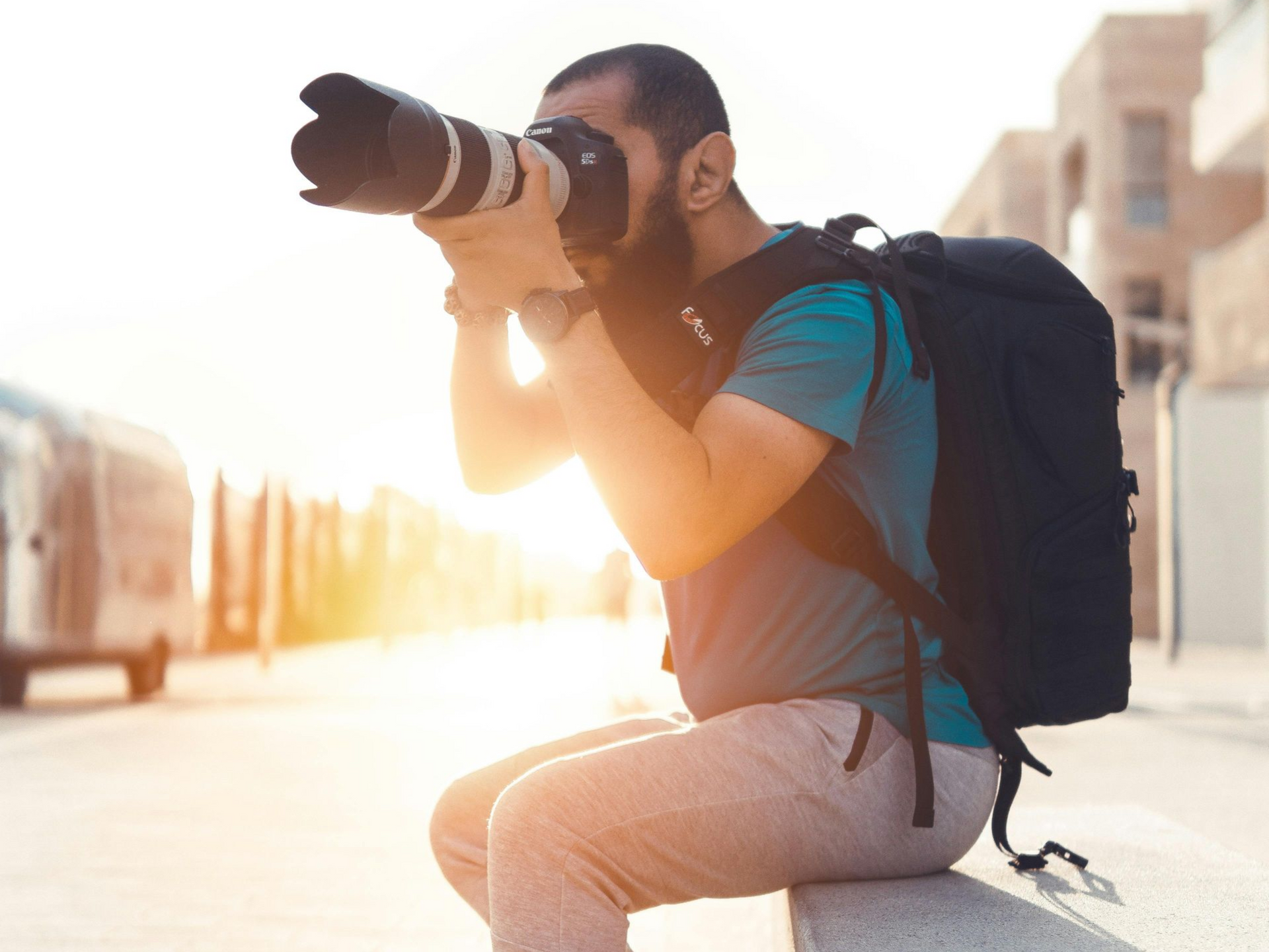Photographe avec appareil photo et sac à dos, assis dehors, prenant une photo, ensoleillé.