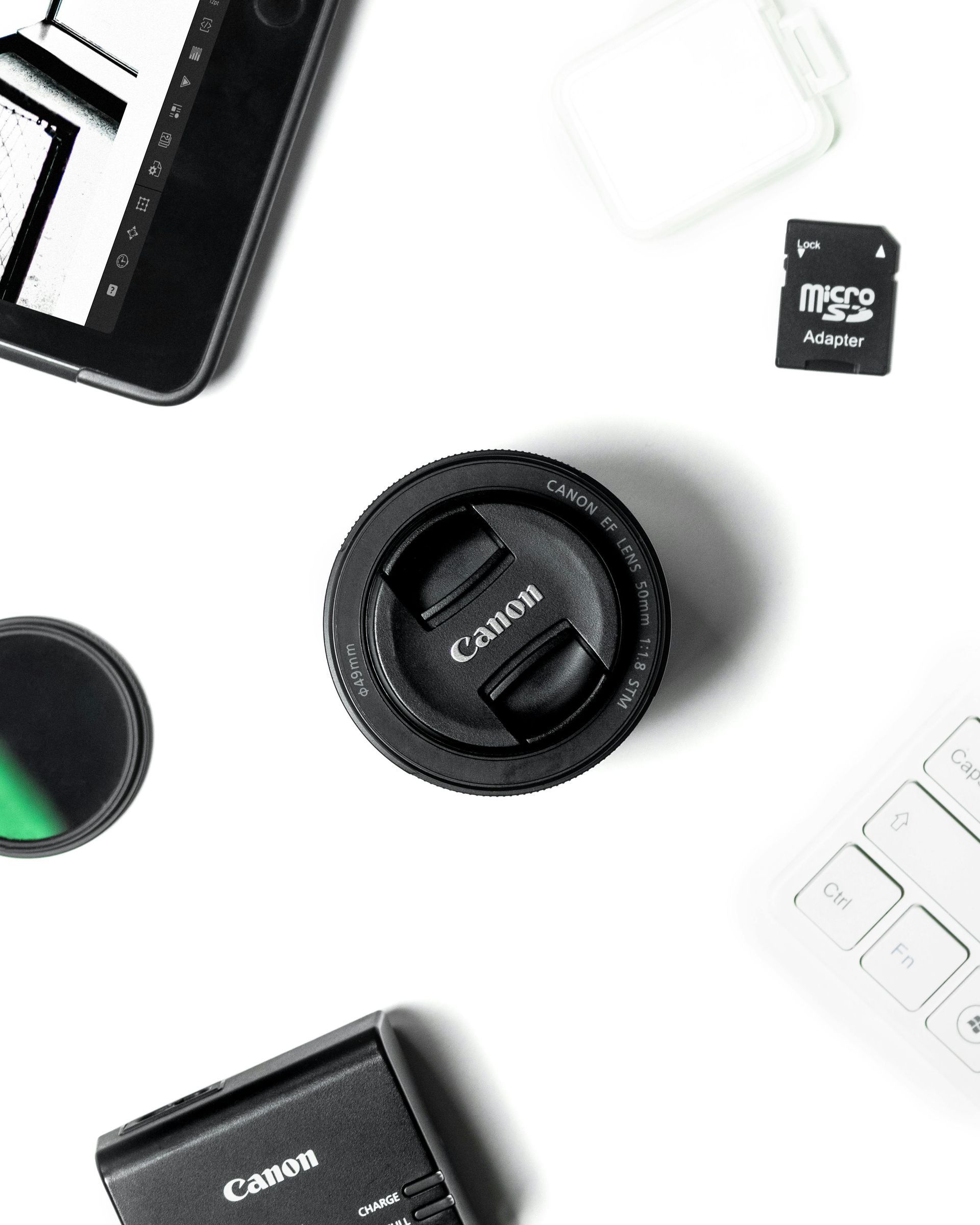 Objectif d'appareil photo entouré d'accessoires technologiques : tablette, carte SD, clavier, chargeur de batterie Canon et filtre.