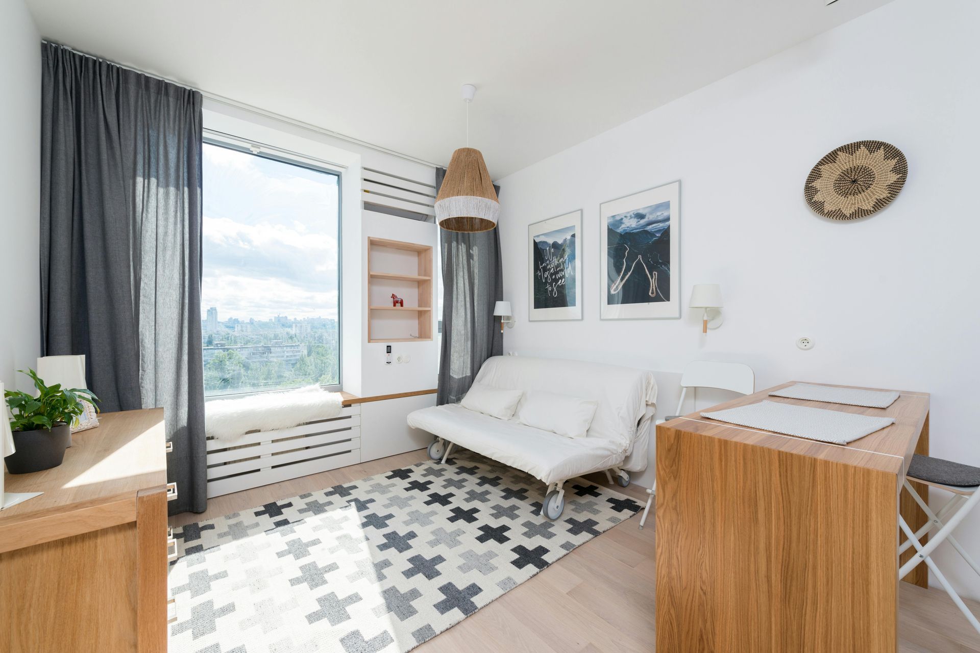 Petit espace de vie lumineux avec un canapé blanc, une fenêtre donnant sur la ville et des meubles en bois.